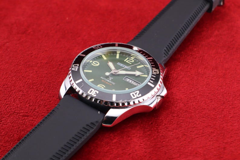 Puede incluir: Un reloj de pulsera de acero inoxidable con bisel negro y correa de caucho negra. La esfera del reloj es verde con n&uacute;meros y agujas blancas. El reloj est&aacute; inclinado sobre una superficie roja.