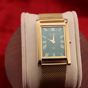 K&ouml;nnte beinhalten: Rechteckige goldfarbene Armbanduhr mit t&uuml;rkisblauem Zifferblatt und goldenem Mesh-Armband. Das Zifferblatt hat r&ouml;mische Ziffern und den Markennamen. Die Uhr wird auf einem wei&szlig;en Kissen vor rotem Hintergrund pr&auml;sentiert.