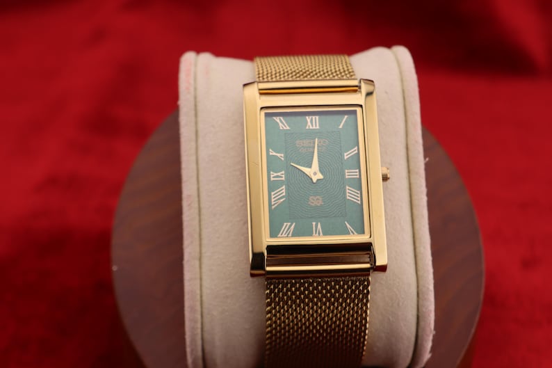K&ouml;nnte beinhalten: Rechteckige goldfarbene Armbanduhr mit t&uuml;rkisblauem Zifferblatt und goldenem Mesh-Armband. Das Zifferblatt hat r&ouml;mische Ziffern und den Markennamen. Die Uhr wird auf einem wei&szlig;en Kissen vor rotem Hintergrund pr&auml;sentiert.