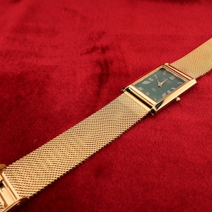 K&ouml;nnte beinhalten: Eine rechteckige Armbanduhr in Gold mit gr&uuml;nem Zifferblatt und r&ouml;mischen Ziffern. Die Uhr hat ein Mesh-Armband und liegt auf rotem Hintergrund.