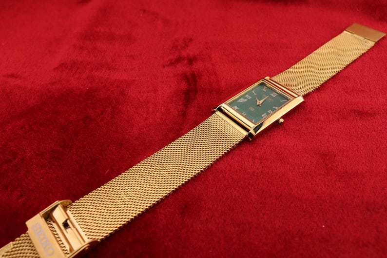 K&ouml;nnte beinhalten: Eine rechteckige Armbanduhr in Gold mit gr&uuml;nem Zifferblatt und r&ouml;mischen Ziffern. Die Uhr hat ein Mesh-Armband und liegt auf rotem Hintergrund.