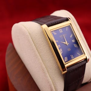May include: A gold rectangular watch with a blue face and Roman numeral hour markers. The watch has gold hands and a brown leather strap. The watch is displayed on a cream cushion against a red background.