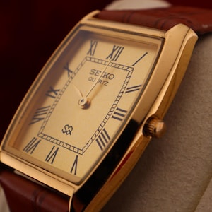 May include: A gold-toned Seiko Quartz watch with a rectangular face and brown leather band. The watch face features black Roman numerals and the Seiko Quartz logo. The watch is set against a neutral background.