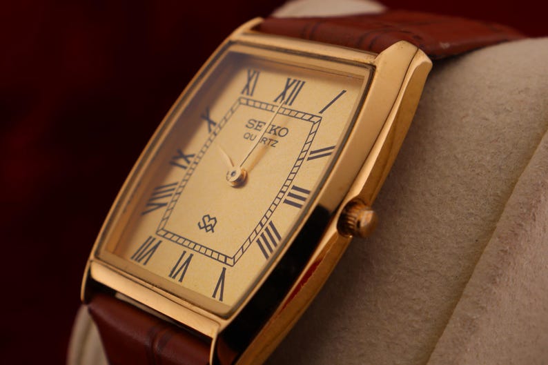 May include: A gold-toned Seiko Quartz watch with a rectangular face and brown leather band. The watch face features black Roman numerals and the Seiko Quartz logo. The watch is set against a neutral background.