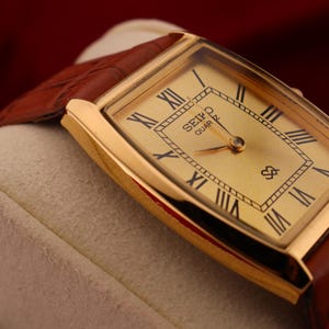 May include: A gold-toned Seiko Quartz watch with a rectangular face and brown leather band. The watch face features black Roman numerals and the Seiko Quartz logo. The watch is displayed on a cream-colored surface.