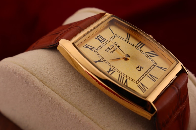 May include: A gold-toned Seiko Quartz watch with a rectangular face and brown leather band. The watch face features black Roman numerals and the Seiko Quartz logo. The watch is displayed on a cream-colored surface.