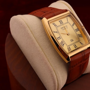 May include: A gold-toned Seiko Quartz watch with a rectangular face and Roman numeral hour markers. The watch has a brown leather band and is displayed on a cream-colored cushion. The watch face displays the Seiko Quartz logo.