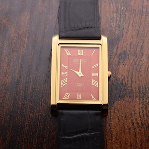 Op de afbeelding: Een goudkleurig rechthoekig Seiko horloge met een rode wijzerplaat en Romeinse cijfers. Het horloge heeft gouden wijzers en een zwarte leren band. Het horloge wordt getoond op een donker houten oppervlak.