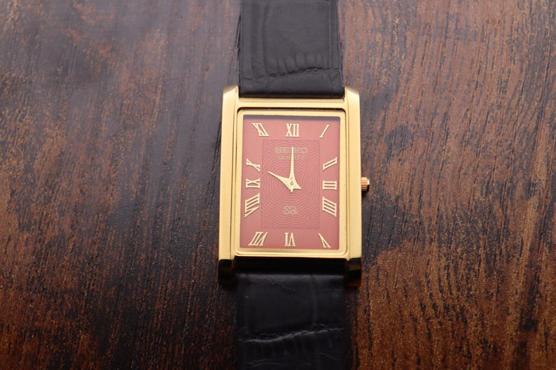 Op de afbeelding: Een goudkleurig rechthoekig Seiko horloge met een rode wijzerplaat en Romeinse cijfers. Het horloge heeft gouden wijzers en een zwarte leren band. Het horloge wordt getoond op een donker houten oppervlak.