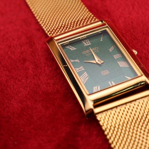 K&ouml;nnte beinhalten: Eine goldfarbene Seiko Quartz-Uhr mit einem rechteckigen gr&uuml;nen Zifferblatt und goldenen r&ouml;mischen Ziffern. Die Uhr hat ein goldenes Mesh-Armband und liegt auf rotem Hintergrund.