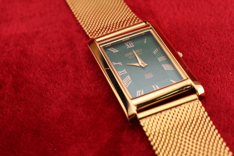 K&ouml;nnte beinhalten: Eine goldfarbene Seiko Quartz-Uhr mit einem rechteckigen gr&uuml;nen Zifferblatt und goldenen r&ouml;mischen Ziffern. Die Uhr hat ein goldenes Mesh-Armband und liegt auf rotem Hintergrund.