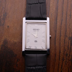 Puede incluir: Un reloj Seiko Quartz rectangular con caja plateada y correa de cuero negro. La esfera del reloj es blanca con números romanos y el nombre de la marca. El reloj se muestra sobre una superficie de madera.