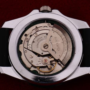 Puede incluir: Primer plano de un reloj plateado con una correa negra, que muestra el intrincado mecanismo interno. La esfera del reloj revela engranajes, resortes y la inscripci&oacute;n "SEIKO JAPAN 63 SEVENTEEN 17 JEWELS". El reloj est&aacute; sobre un fondo rojo oscuro.