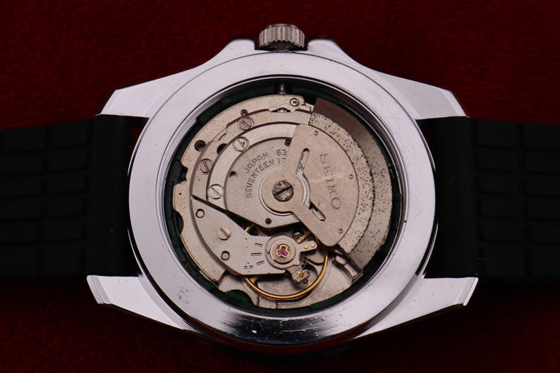 Puede incluir: Primer plano de un reloj plateado con una correa negra, que muestra el intrincado mecanismo interno. La esfera del reloj revela engranajes, resortes y la inscripci&oacute;n "SEIKO JAPAN 63 SEVENTEEN 17 JEWELS". El reloj est&aacute; sobre un fondo rojo oscuro.