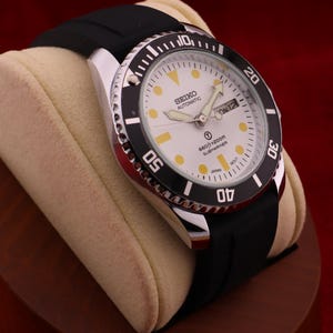May include: A Seiko Automatic Submariner watch with a white dial, black bezel, and black rubber strap. The watch features a day and date display and is displayed on a beige watch pillow.
