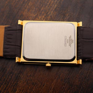 Op de afbeelding: Een goudkleurig rechthoekig horloge met een zilveren wijzerplaat en een bruine leren band. De band is donkerbruin bij de wijzerplaat en lichtbruin aan de buitenranden. Het horloge ligt op een donkerbruin houten oppervlak.