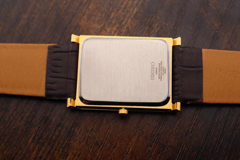 Op de afbeelding: Een goudkleurig rechthoekig horloge met een zilveren wijzerplaat en een bruine leren band. De band is donkerbruin bij de wijzerplaat en lichtbruin aan de buitenranden. Het horloge ligt op een donkerbruin houten oppervlak.