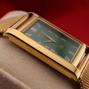 K&ouml;nnte beinhalten: Eine rechteckige Armbanduhr in Gold mit gr&uuml;nem Zifferblatt und goldenen Zeigern. Die Uhr hat r&ouml;mische Ziffern als Stundenmarkierungen und ein goldenes Mesh-Armband. Die Uhr wird vor einem neutralen Hintergrund mit rotem Hintergrund gezeigt.