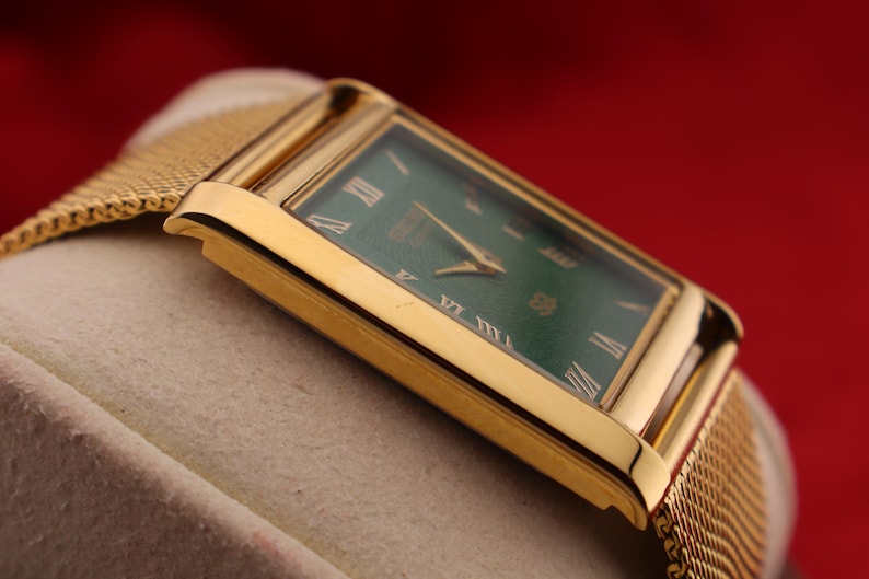 K&ouml;nnte beinhalten: Eine rechteckige Armbanduhr in Gold mit gr&uuml;nem Zifferblatt und goldenen Zeigern. Die Uhr hat r&ouml;mische Ziffern als Stundenmarkierungen und ein goldenes Mesh-Armband. Die Uhr wird vor einem neutralen Hintergrund mit rotem Hintergrund gezeigt.