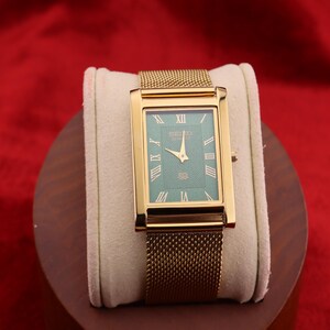 K&ouml;nnte beinhalten: Rechteckige Armbanduhr in Gold mit gr&uuml;nem Zifferblatt und goldenen Zeigern. Die Uhr hat r&ouml;mische Ziffern und ein Mesh-Armband. Die Uhr wird auf einem cremefarbenen Kissen vor rotem Hintergrund pr&auml;sentiert.