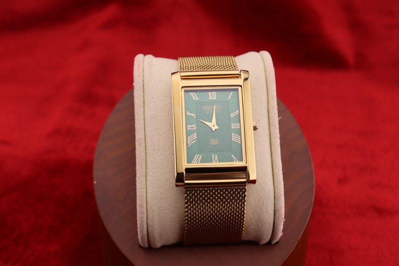 K&ouml;nnte beinhalten: Rechteckige Armbanduhr in Gold mit gr&uuml;nem Zifferblatt und goldenen Zeigern. Die Uhr hat r&ouml;mische Ziffern und ein Mesh-Armband. Die Uhr wird auf einem cremefarbenen Kissen vor rotem Hintergrund pr&auml;sentiert.
