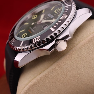 Puede incluir: Primer plano de un reloj autom&aacute;tico Seiko con bisel negro y correa de caucho negra. La esfera del reloj es verde oscuro con agujas e indicadores luminosos. El reloj est&aacute; sobre un fondo beige.