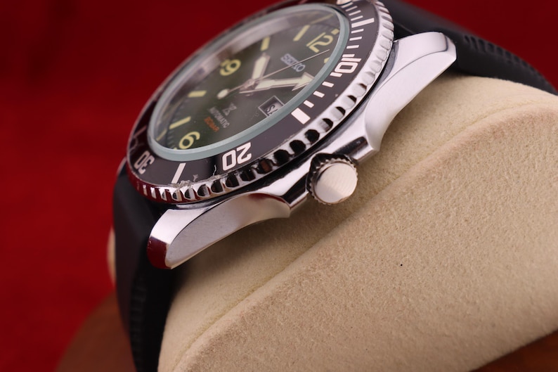 Puede incluir: Primer plano de un reloj autom&aacute;tico Seiko con bisel negro y correa de caucho negra. La esfera del reloj es verde oscuro con agujas e indicadores luminosos. El reloj est&aacute; sobre un fondo beige.