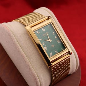 K&ouml;nnte beinhalten: Eine rechteckige goldfarbene Armbanduhr mit gr&uuml;nem Zifferblatt und goldenem Mesh-Armband. Die Uhr hat r&ouml;mische Ziffern und goldene Zeiger. Die Uhr wird auf einem cremefarbenen Kissen vor rotem Hintergrund pr&auml;sentiert.