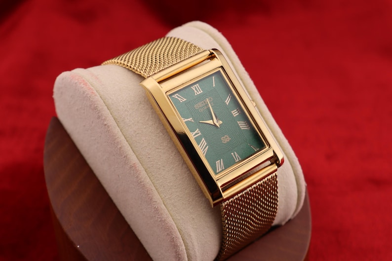 K&ouml;nnte beinhalten: Eine rechteckige goldfarbene Armbanduhr mit gr&uuml;nem Zifferblatt und goldenem Mesh-Armband. Die Uhr hat r&ouml;mische Ziffern und goldene Zeiger. Die Uhr wird auf einem cremefarbenen Kissen vor rotem Hintergrund pr&auml;sentiert.