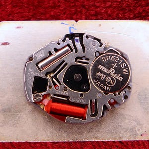 May include: Close-up of a watch movement with a silver-colored metal base. The movement features a battery labeled "SR621SW" and components with the text "JAPAN". A red coil and other small parts are visible.