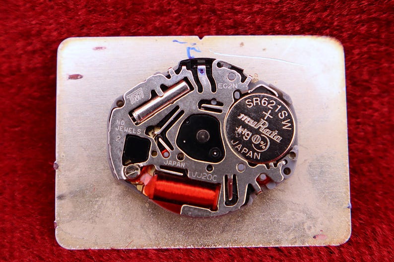May include: Close-up of a watch movement with a silver-colored metal base. The movement features a battery labeled "SR621SW" and components with the text "JAPAN". A red coil and other small parts are visible.