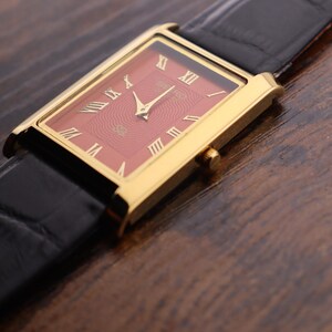 Op de afbeelding: Een gouden rechthoekig polshorloge met een rode wijzerplaat en gouden Romeinse cijfers. Het horloge heeft gouden wijzers en een zwarte leren band. Het horloge staat tegen een donkere houtnerf achtergrond.