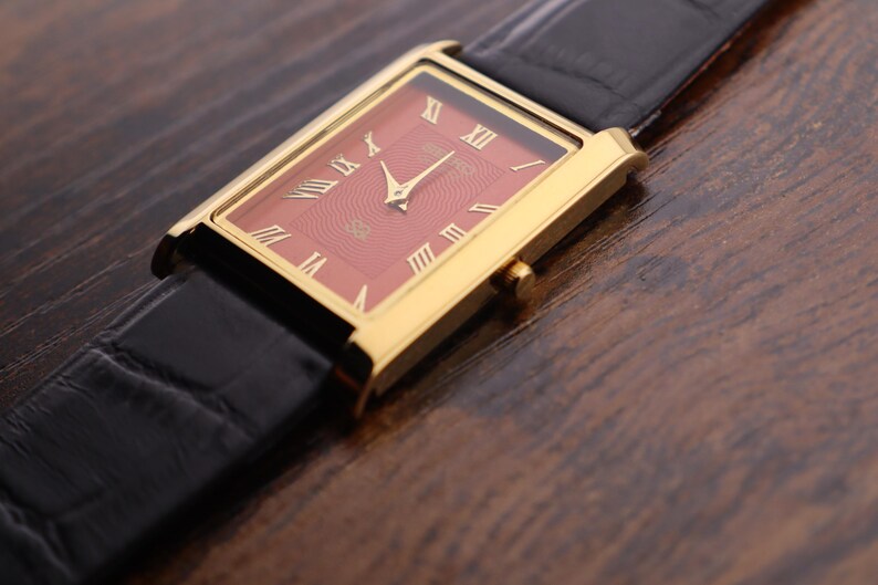 Op de afbeelding: Een gouden rechthoekig polshorloge met een rode wijzerplaat en gouden Romeinse cijfers. Het horloge heeft gouden wijzers en een zwarte leren band. Het horloge staat tegen een donkere houtnerf achtergrond.