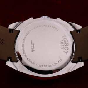 Puede incluir: Primer plano de un reloj Tissot de acero inoxidable plateado. La parte trasera del reloj muestra "TISSOT 1853" y es resistente al agua hasta 100m. Correa de cuero marr&oacute;n y cristal de zafiro.