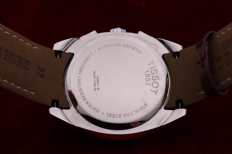 Puede incluir: Primer plano de un reloj Tissot de acero inoxidable plateado. La parte trasera del reloj muestra "TISSOT 1853" y es resistente al agua hasta 100m. Correa de cuero marr&oacute;n y cristal de zafiro.