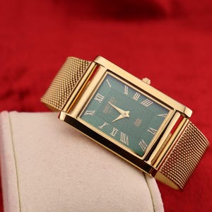 K&ouml;nnte beinhalten: Eine rechteckige goldfarbene Armbanduhr mit gr&uuml;nem Zifferblatt und r&ouml;mischen Ziffern. Die Uhr hat ein Mesh-Armband und wird auf einer wei&szlig;en Oberfl&auml;che vor rotem Hintergrund pr&auml;sentiert.