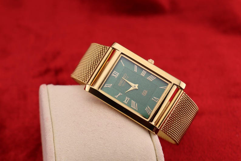 K&ouml;nnte beinhalten: Eine rechteckige goldfarbene Armbanduhr mit gr&uuml;nem Zifferblatt und r&ouml;mischen Ziffern. Die Uhr hat ein Mesh-Armband und wird auf einer wei&szlig;en Oberfl&auml;che vor rotem Hintergrund pr&auml;sentiert.