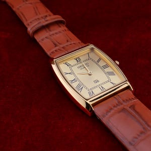 May include: A gold-toned Seiko Quartz watch with a rectangular face and Roman numeral hour markers. The watch has a brown leather band with a crocodile pattern. The watch is displayed on a dark red surface.