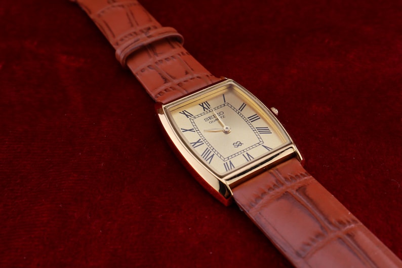 May include: A gold-toned Seiko Quartz watch with a rectangular face and Roman numeral hour markers. The watch has a brown leather band with a crocodile pattern. The watch is displayed on a dark red surface.