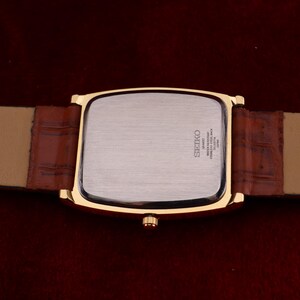 May include: A gold-toned rectangular Seiko watch with a brown leather band. The watch face is silver, and the band is a lighter brown. The back of the watch has the Seiko logo and other text. The watch band is 20 cm long.