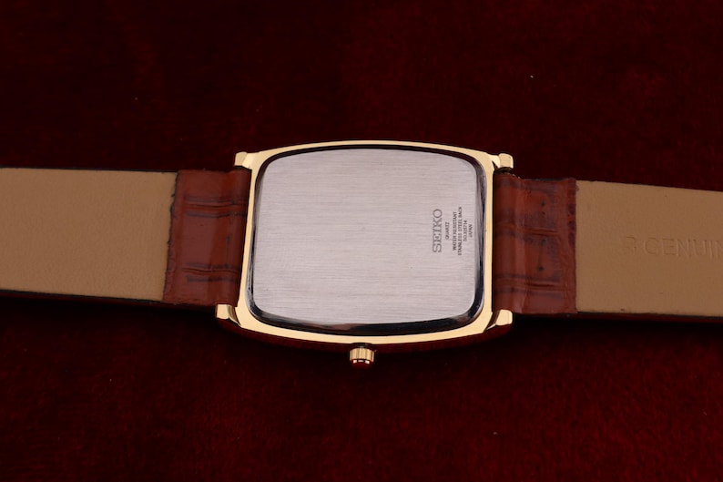 May include: A gold-toned rectangular Seiko watch with a brown leather band. The watch face is silver, and the band is a lighter brown. The back of the watch has the Seiko logo and other text. The watch band is 20 cm long.