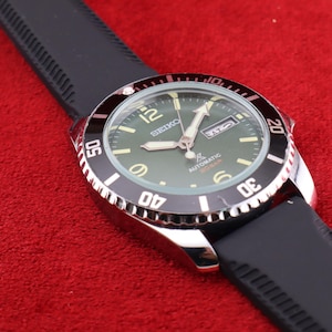 Puede incluir: Un reloj de pulsera autom&aacute;tico Seiko con una correa de goma negra. El reloj tiene una esfera verde con manecillas y marcadores blancos y plateados. El bisel es negro y plateado, con n&uacute;meros que indican los minutos. El reloj est&aacute; sobre una superficie roja.