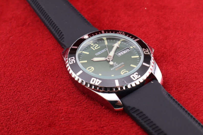 Puede incluir: Un reloj de pulsera autom&aacute;tico Seiko con una correa de goma negra. El reloj tiene una esfera verde con manecillas y marcadores blancos y plateados. El bisel es negro y plateado, con n&uacute;meros que indican los minutos. El reloj est&aacute; sobre una superficie roja.