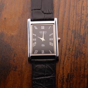 May include: A rectangular Seiko Quartz watch with a black face and silver-colored hands and numerals. The watch has a black leather band and a silver-colored case. The watch is displayed on a brown wooden surface.