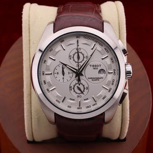 Puede incluir: Un reloj cron&oacute;grafo Tissot plateado con esfera clara y correa de cuero marr&oacute;n. El reloj presenta m&uacute;ltiples subesferas y el texto "Tissot 1853 Chronograph Quartz". El reloj se exhibe sobre un coj&iacute;n color crema.