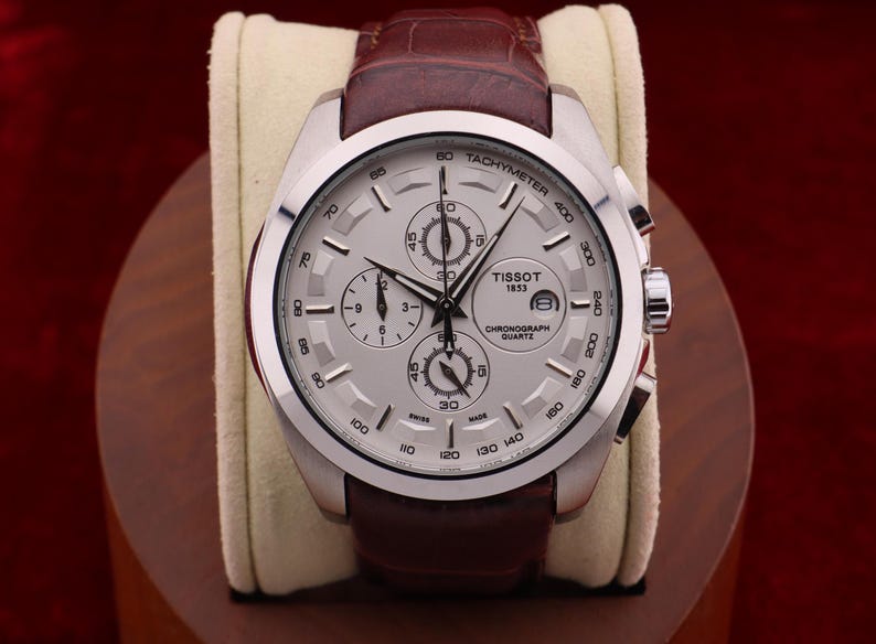 Puede incluir: Un reloj cron&oacute;grafo Tissot plateado con esfera clara y correa de cuero marr&oacute;n. El reloj presenta m&uacute;ltiples subesferas y el texto "Tissot 1853 Chronograph Quartz". El reloj se exhibe sobre un coj&iacute;n color crema.