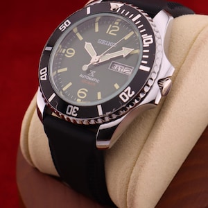 Puede incluir: Un reloj autom&aacute;tico Seiko con bisel negro y correa de caucho negra. La esfera del reloj es verde oscuro con agujas e indicadores luminosos. El reloj muestra el d&iacute;a y la fecha. El reloj se exhibe sobre una almohada beige.