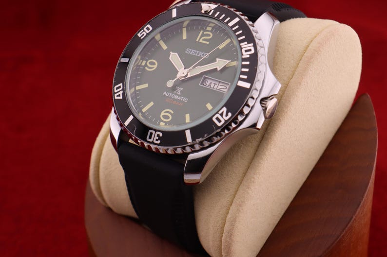 Puede incluir: Un reloj autom&aacute;tico Seiko con bisel negro y correa de caucho negra. La esfera del reloj es verde oscuro con agujas e indicadores luminosos. El reloj muestra el d&iacute;a y la fecha. El reloj se exhibe sobre una almohada beige.