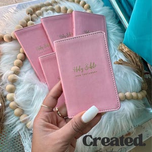 May include: Four pink leather-bound New Testament Holy Bibles with gold lettering. The Bibles are stacked on top of each other. The text "Created with Purpose" is visible in the bottom right corner of the image.