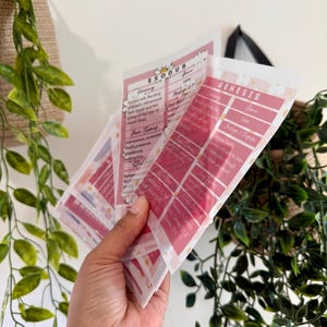 May include: A hand holds several pink and white study cards with text. The cards have a floral design and include the words "EXODUS" and "GENESIS". The cards are surrounded by green leafy plants.